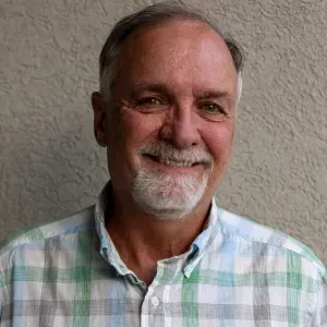 Man with gray hair and beard smiles at camera; wears a light green plaid shirt.