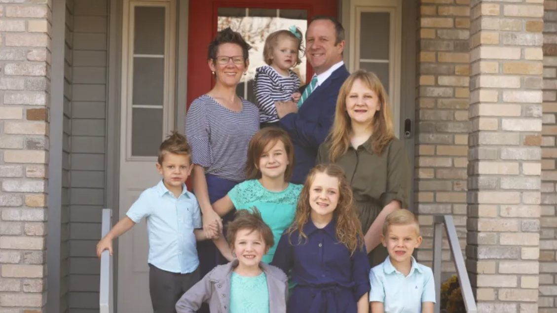 Family of nine poses on porch: parents holding toddler, other children smiling.