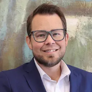 Man in glasses, blue blazer, and white shirt smiling, in front of abstract art.