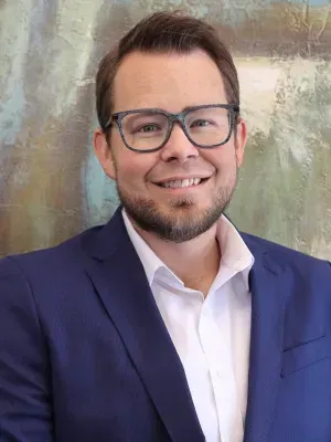 Man in glasses and blue blazer smiling, in front of a blurry background.