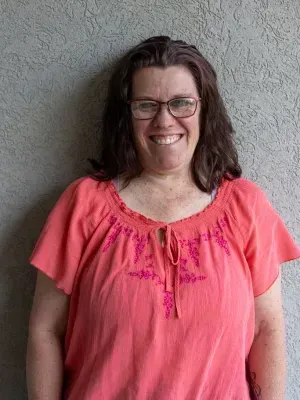 Woman with glasses, pink shirt, smiling in front of a textured grey wall.