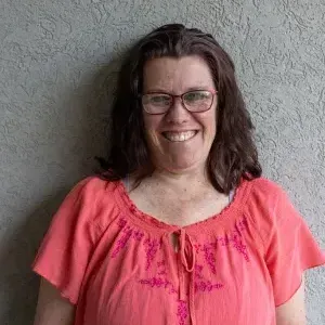 Woman with glasses smiles, wearing coral shirt with pink embroidery against a gray stucco wall.