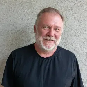 Man with a gray beard wearing a black shirt smiling, in front of a textured wall.