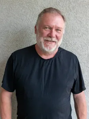 Man with a white beard and black shirt smiles in front of a textured wall.