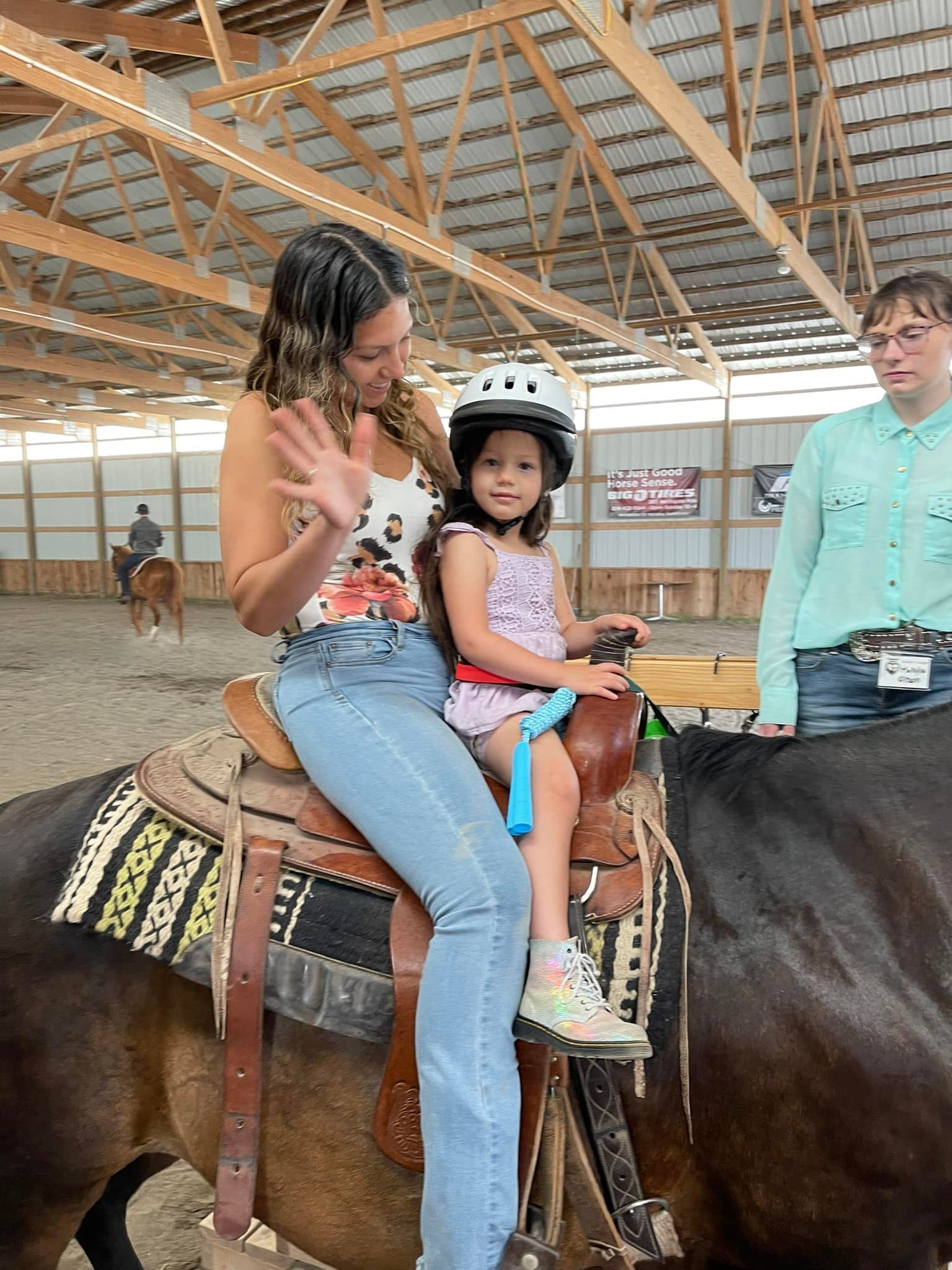 Woman and girl on horseback in an arena. Woman waves. Girl wears a helmet and is harnessed.