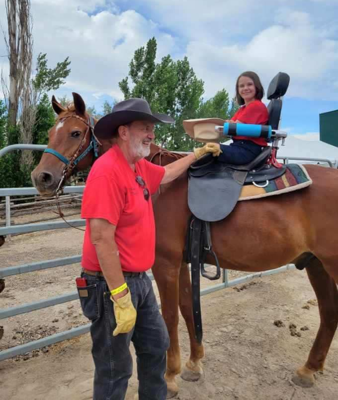 A girl with a disability sits on a horse with support, guided by a man in a red shirt.