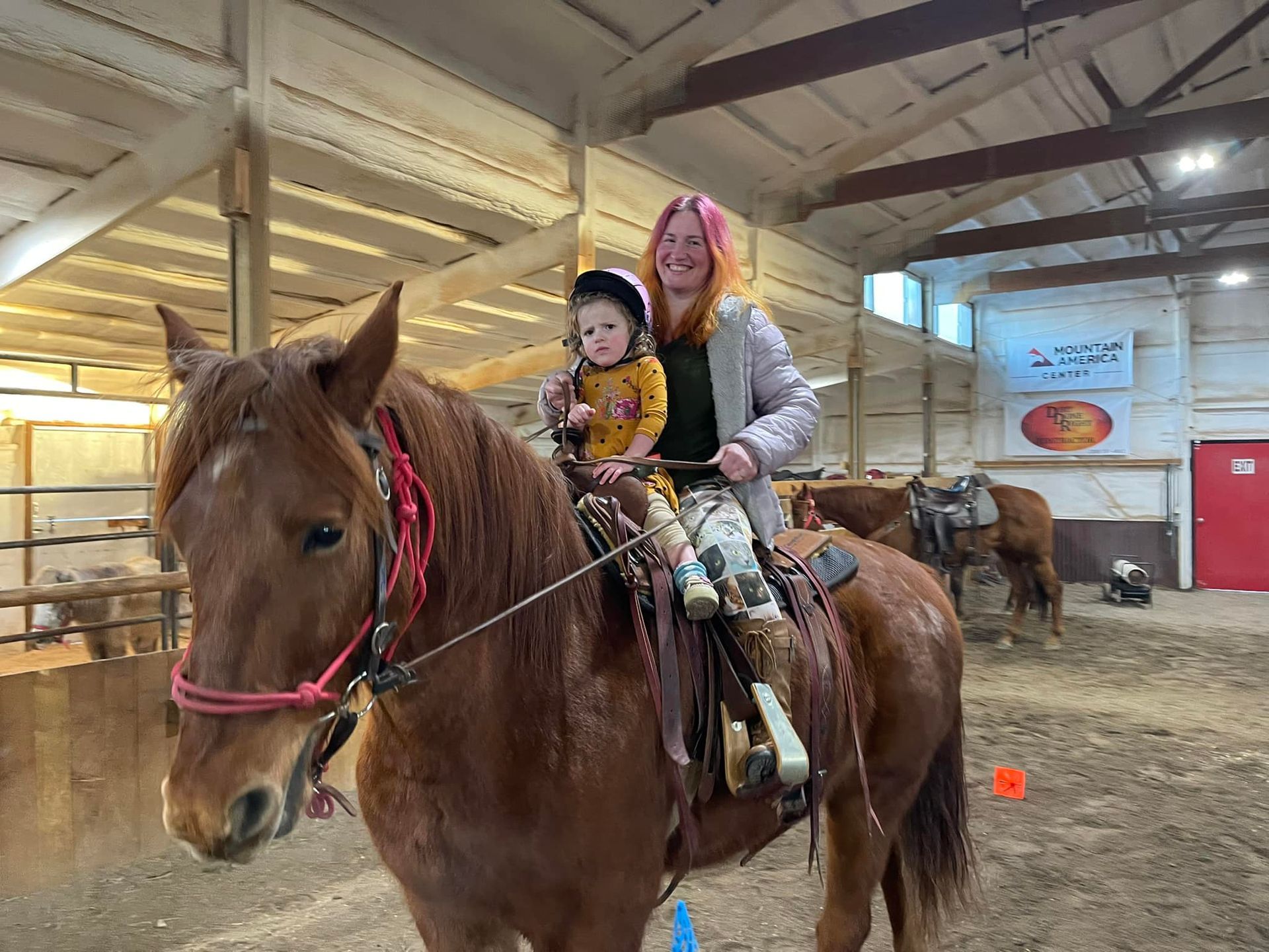Woman and child riding a brown horse inside an indoor riding arena.