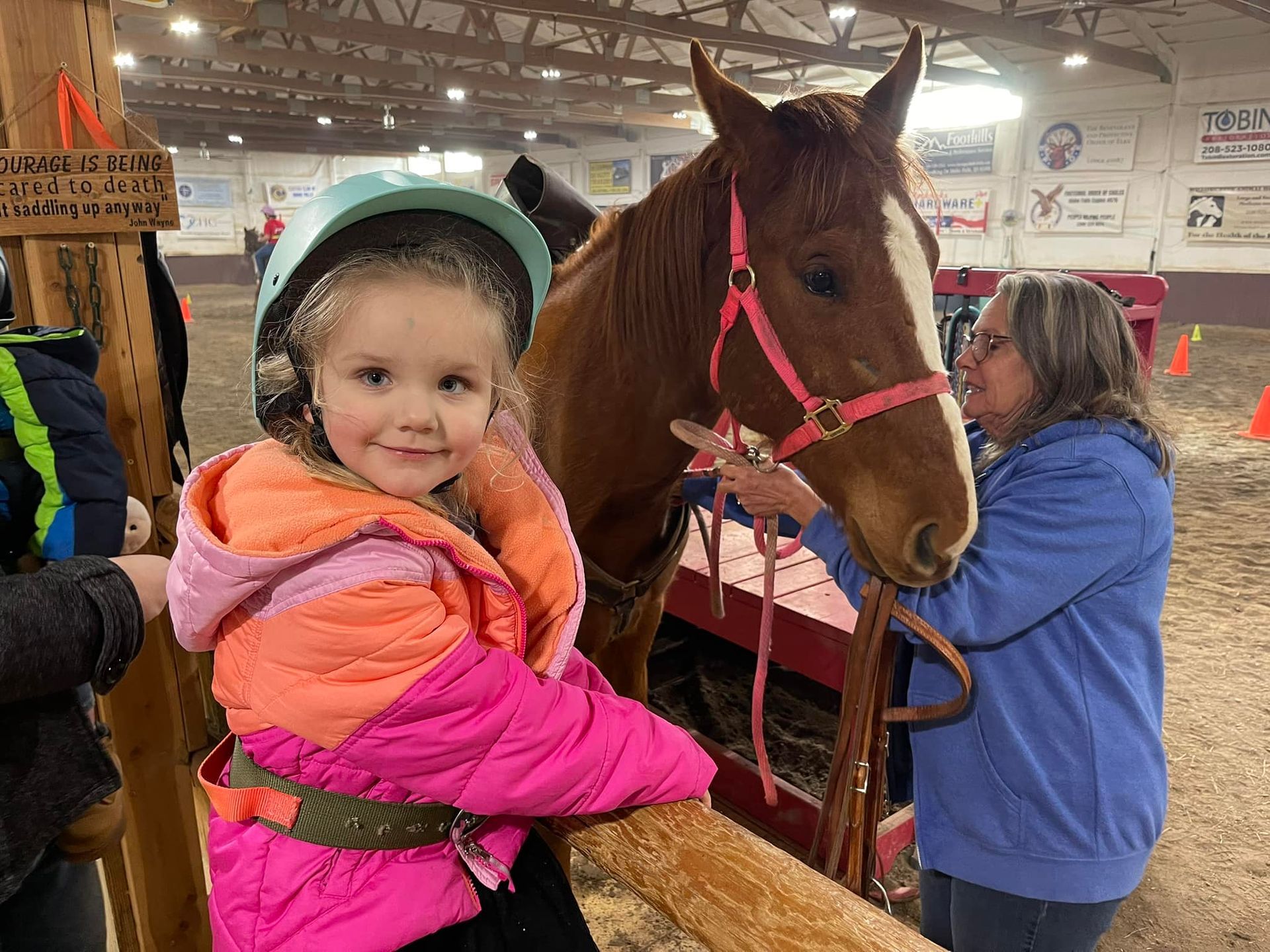 Young girl in helmet smiles beside a brown horse with a pink halter indoors. Woman adjusts the halter.