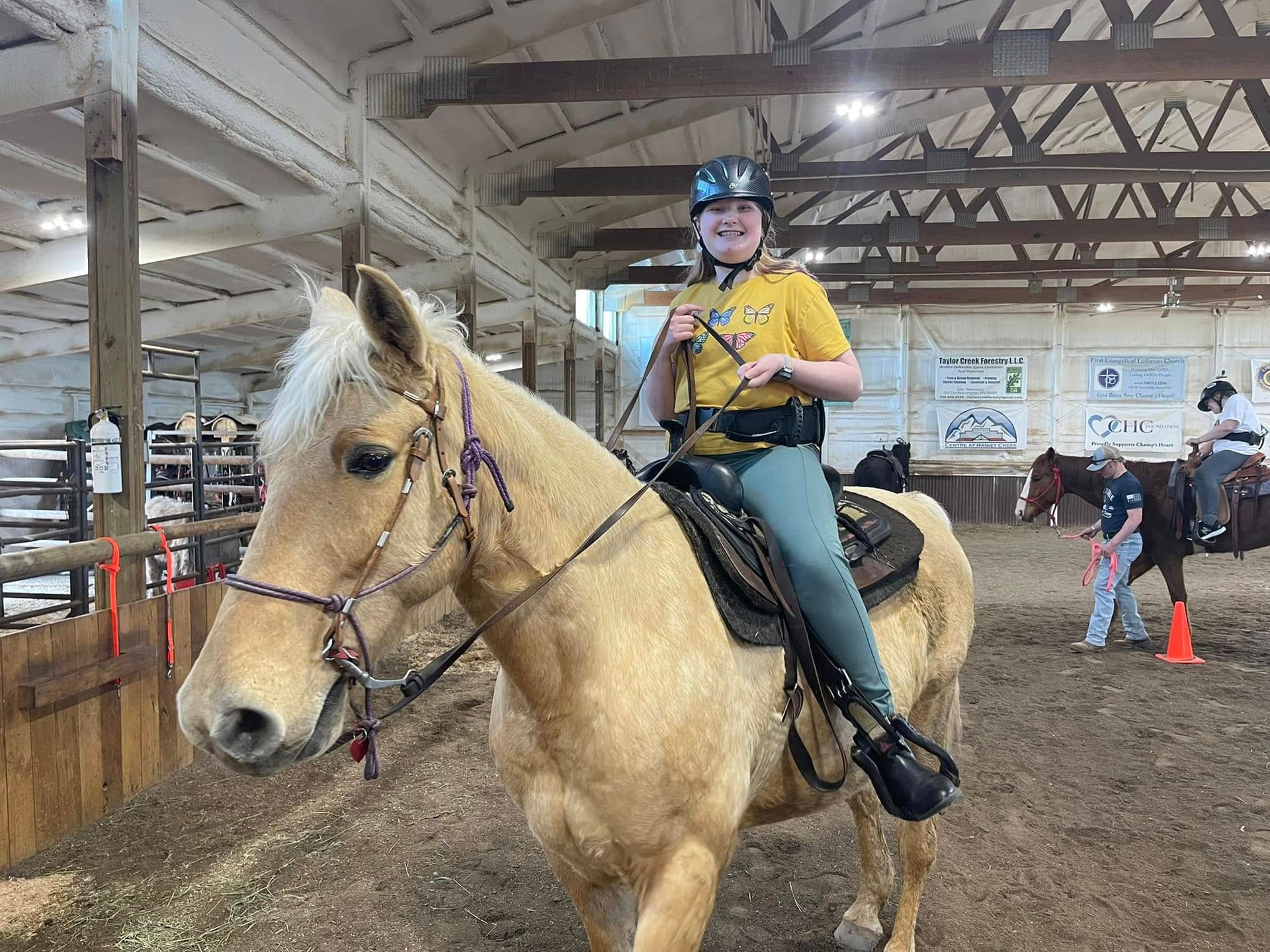Woman on a tan horse, inside riding arena, smiling. She wears a helmet and safety vest.