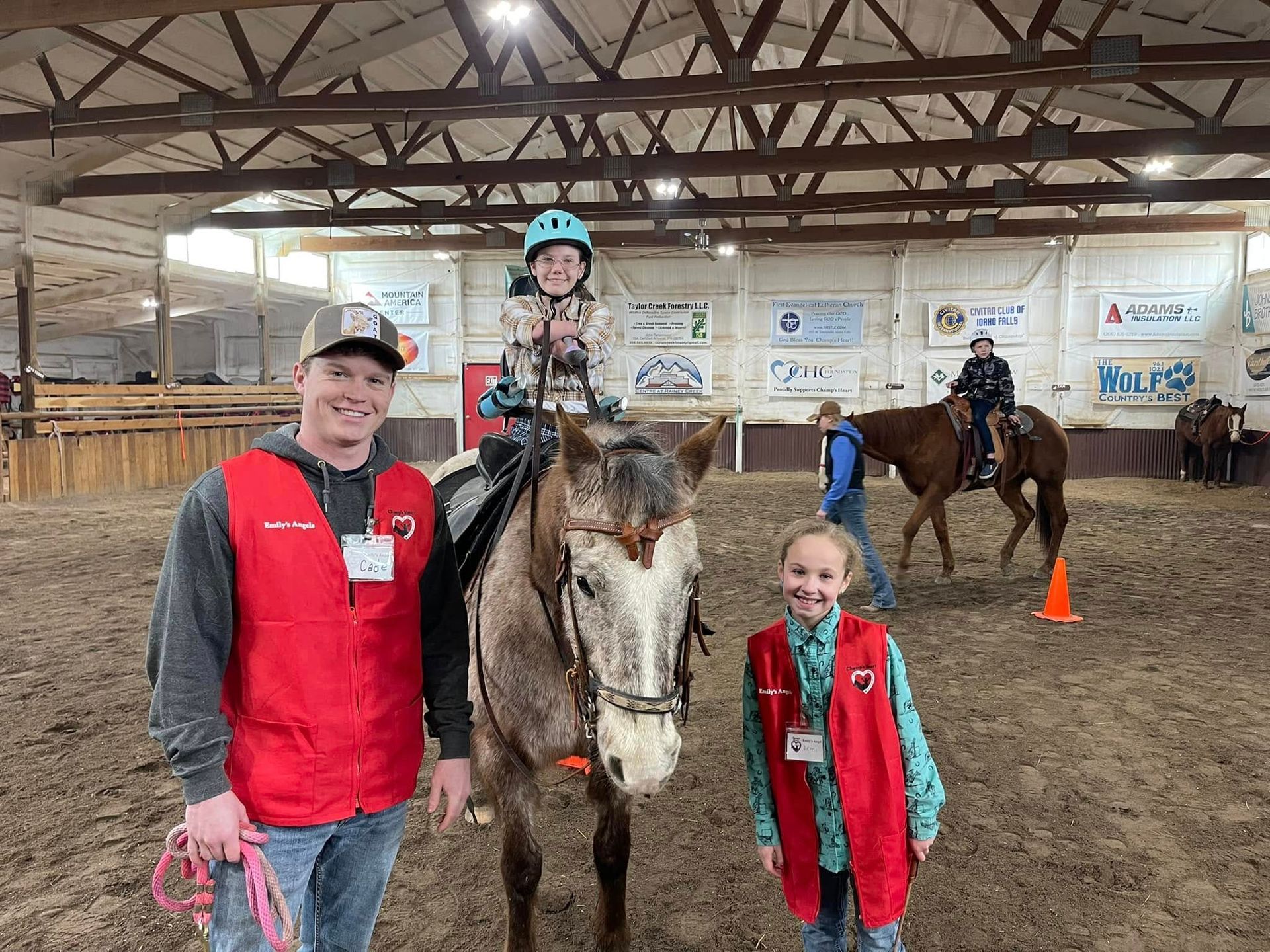 Smiling volunteers and a child on horseback in an indoor riding arena. Another rider in background.