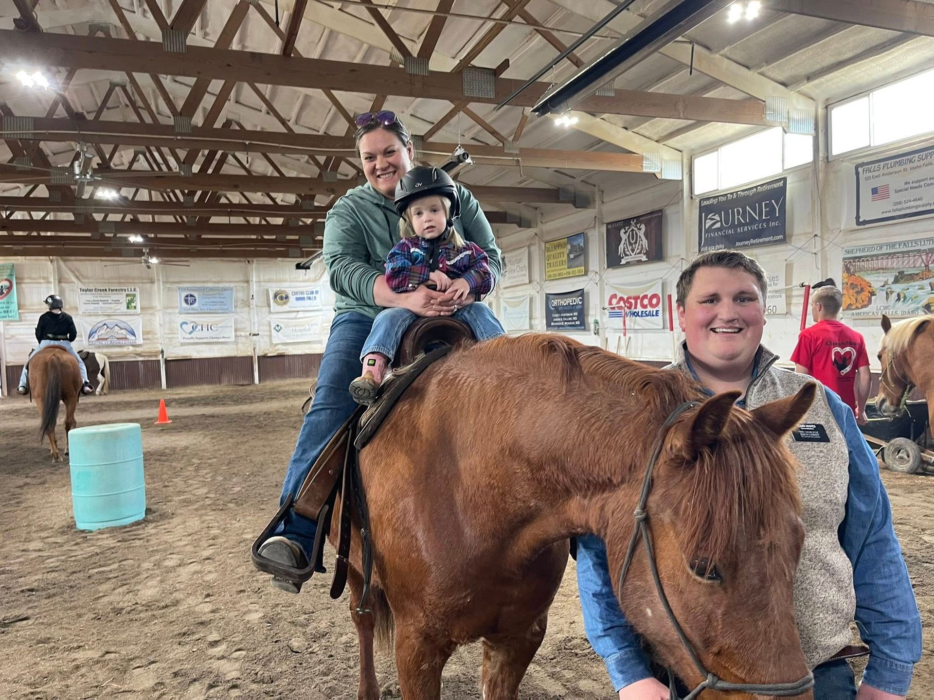 Woman and child on a horse; man smiles beside them in an indoor arena.