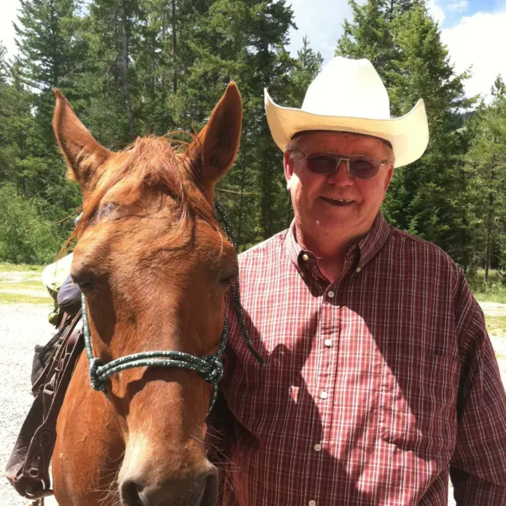 Man in cowboy hat and red plaid shirt smiles next to a brown horse, both outside near trees.