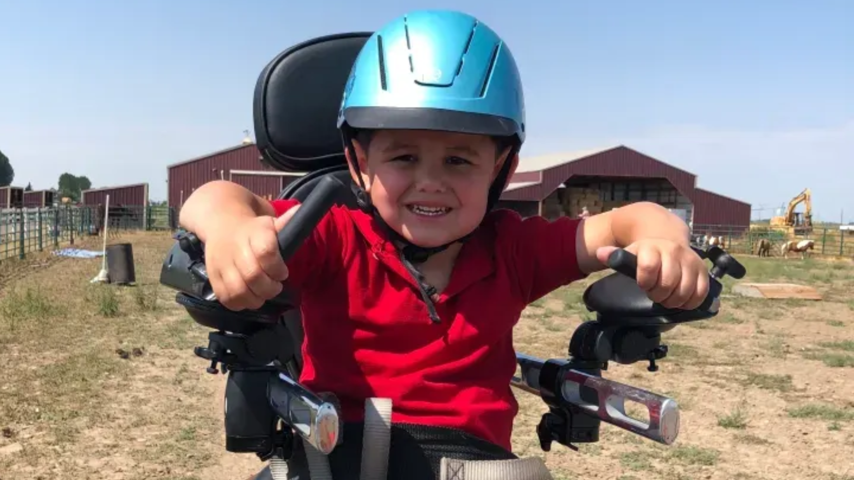 Young boy with a blue helmet, smiling, driving a powered wheelchair outdoors. Red shirt. Barn in background.