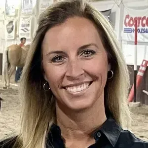 Blond woman smiles, wearing a black shirt. In a barn-like setting with horses and a Costco sign.