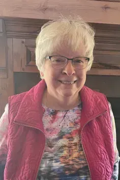 Elderly woman with short white hair, glasses, and red vest, smiles indoors.