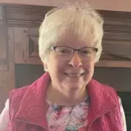 Elderly woman with glasses smiles, wearing a pink vest, in front of a wood panel.
