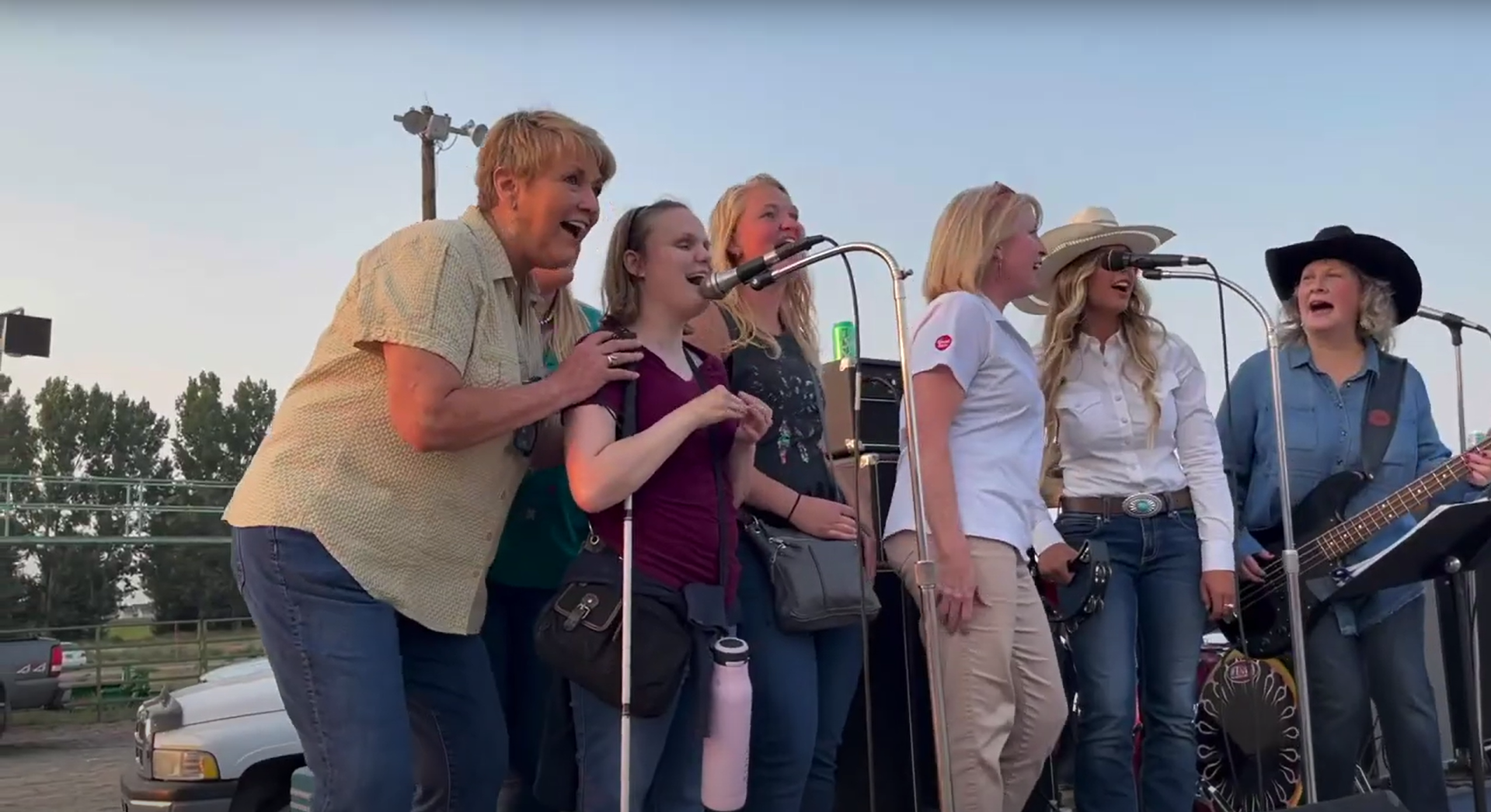 Group of women singing on stage at an outdoor event. Some wear cowboy hats, one plays a guitar.