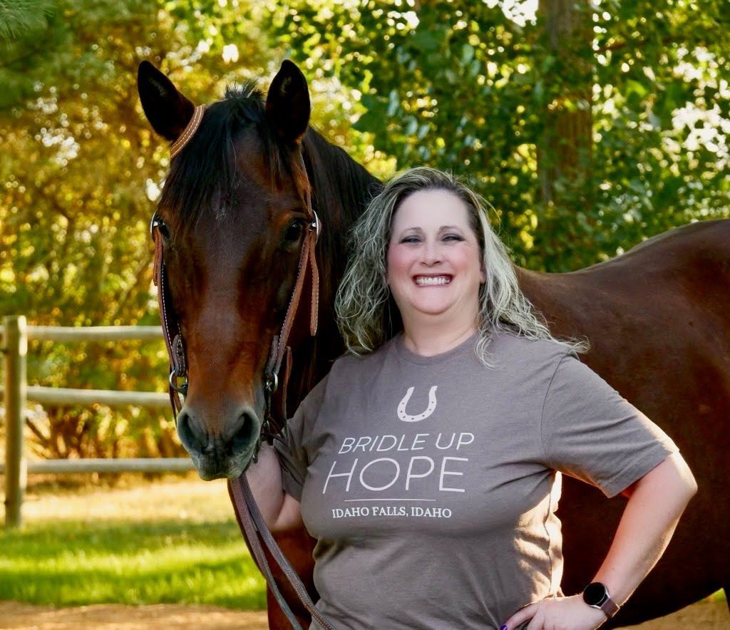 Woman smiles next to brown horse, wearing a t-shirt. Outdoors, sunny setting.