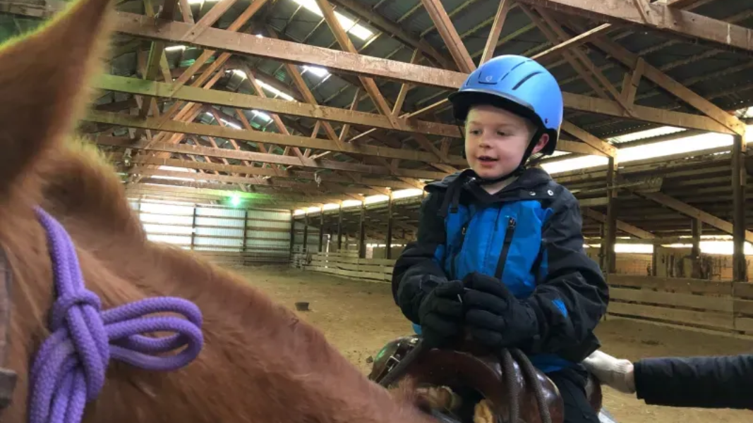 Young child in a blue helmet and jacket rides a brown horse in a stable.