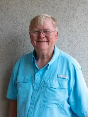 Elderly man with glasses smiles, wearing a blue shirt, against a textured wall.