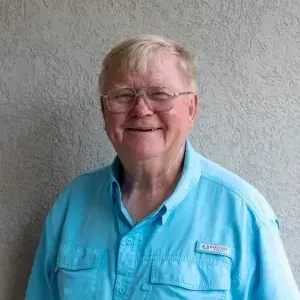 Elderly man with glasses, smiling, wearing a blue button-down shirt. Standing in front of a textured wall.