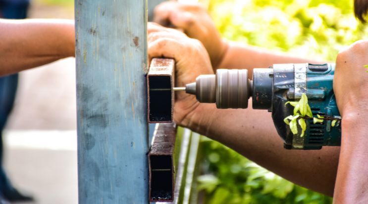 Person using a drill to make a hole in a metal pole. Another person holds the pole steady.