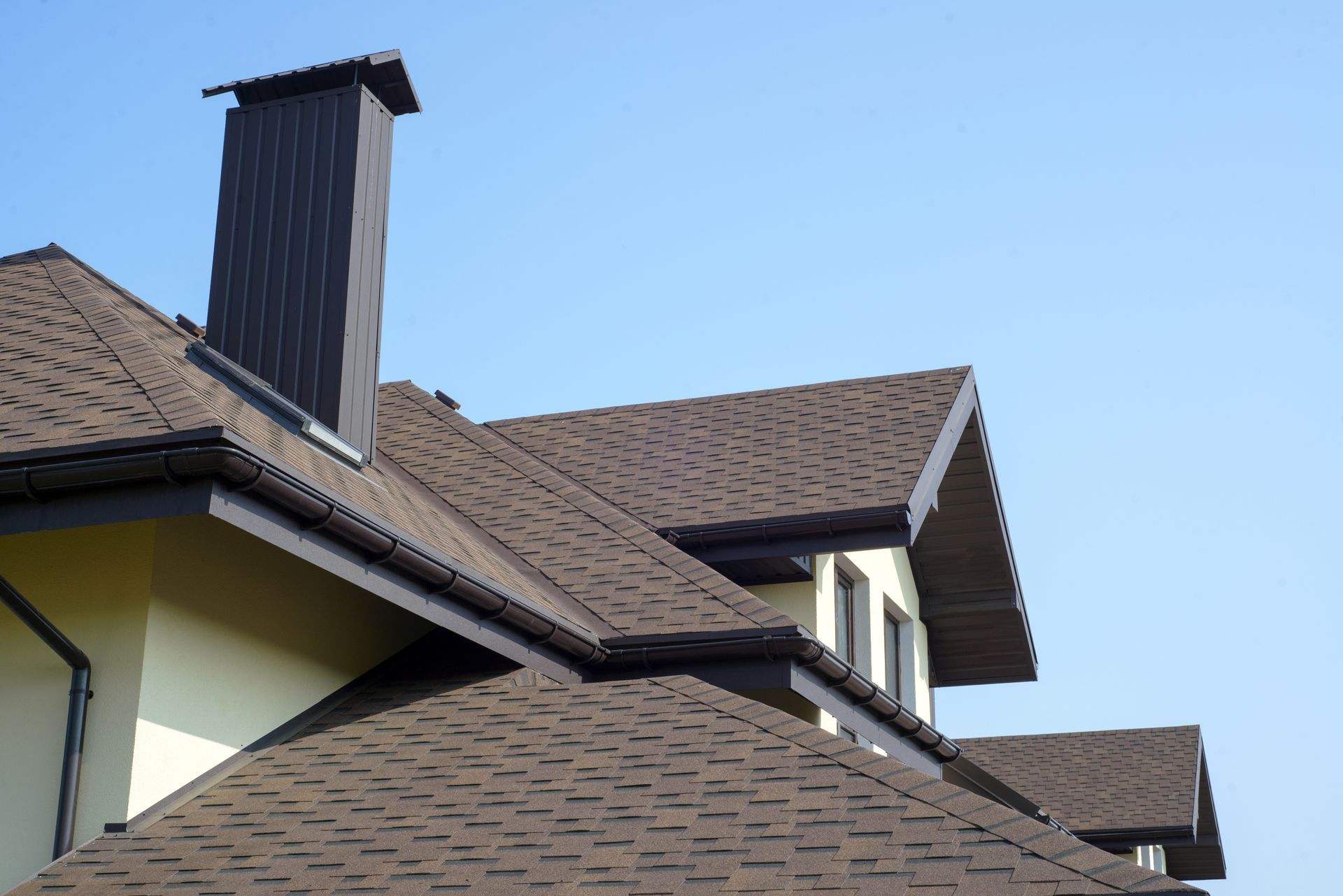 Brown shingle roof with chimney against a blue sky.