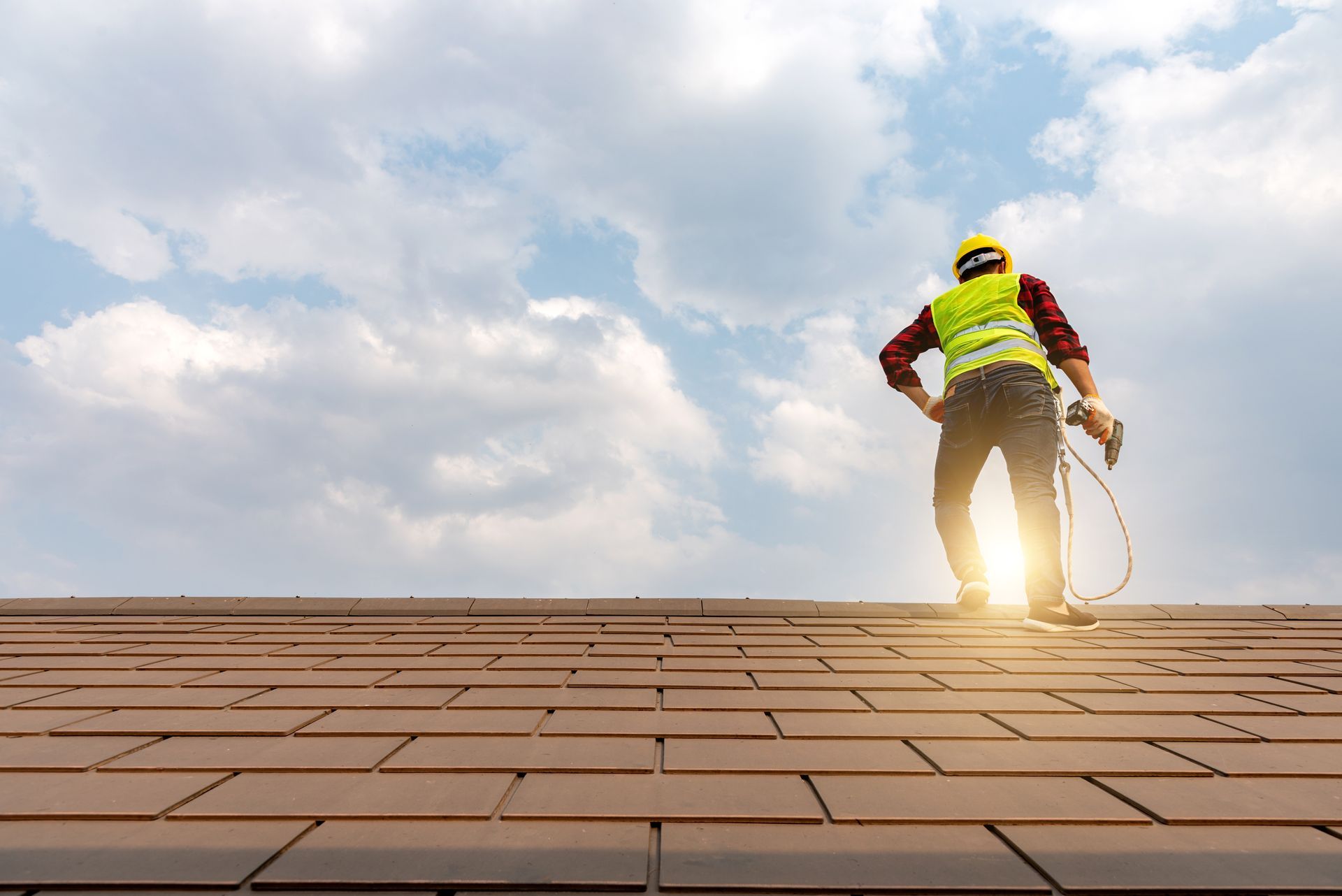 Roofer on a brown tile roof, wearing a hard hat and safety vest, working under a cloudy sky.