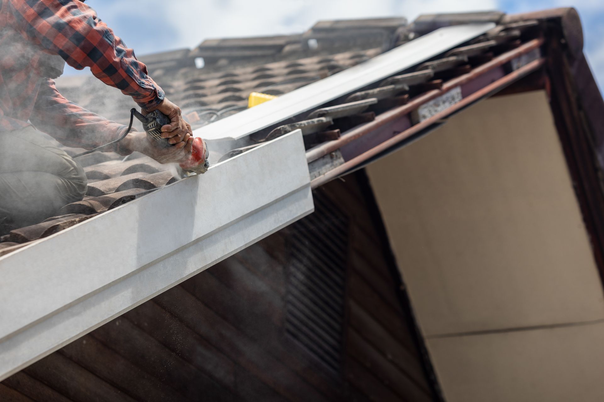 Roofer cutting white gutter with a power saw on a roof, creating dust.