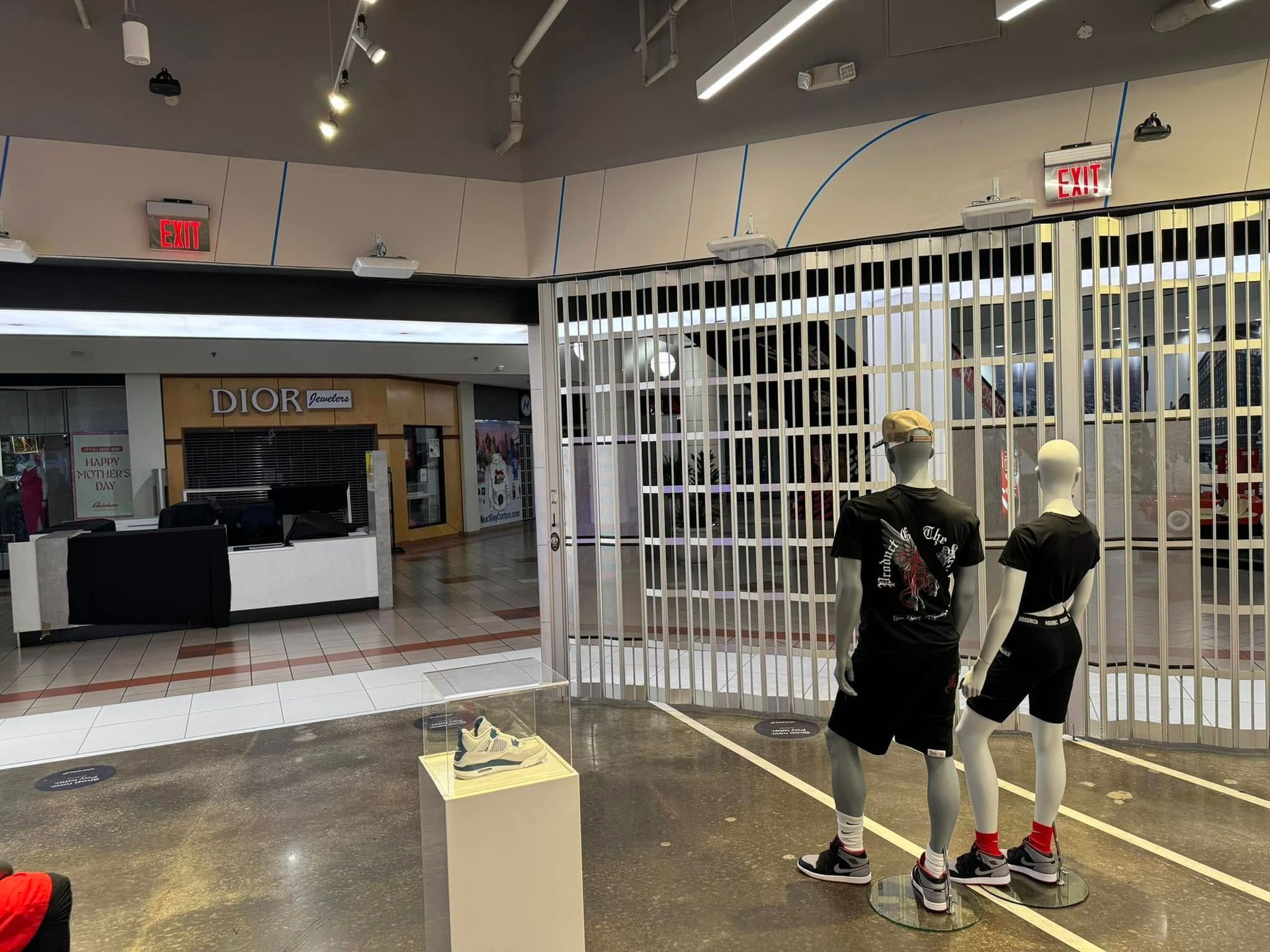 Storefront closed with metal security gate, mannequins wearing black, Dior store in the distance.