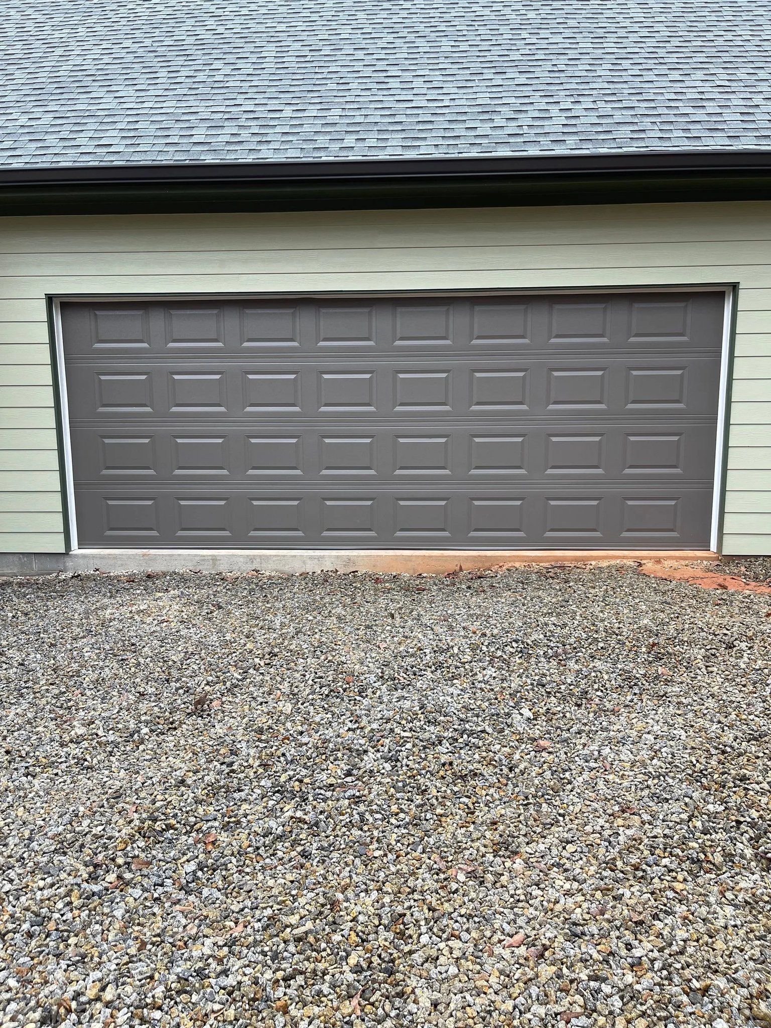 Gray garage door on a light green building, gravel in foreground, shingles on roof.