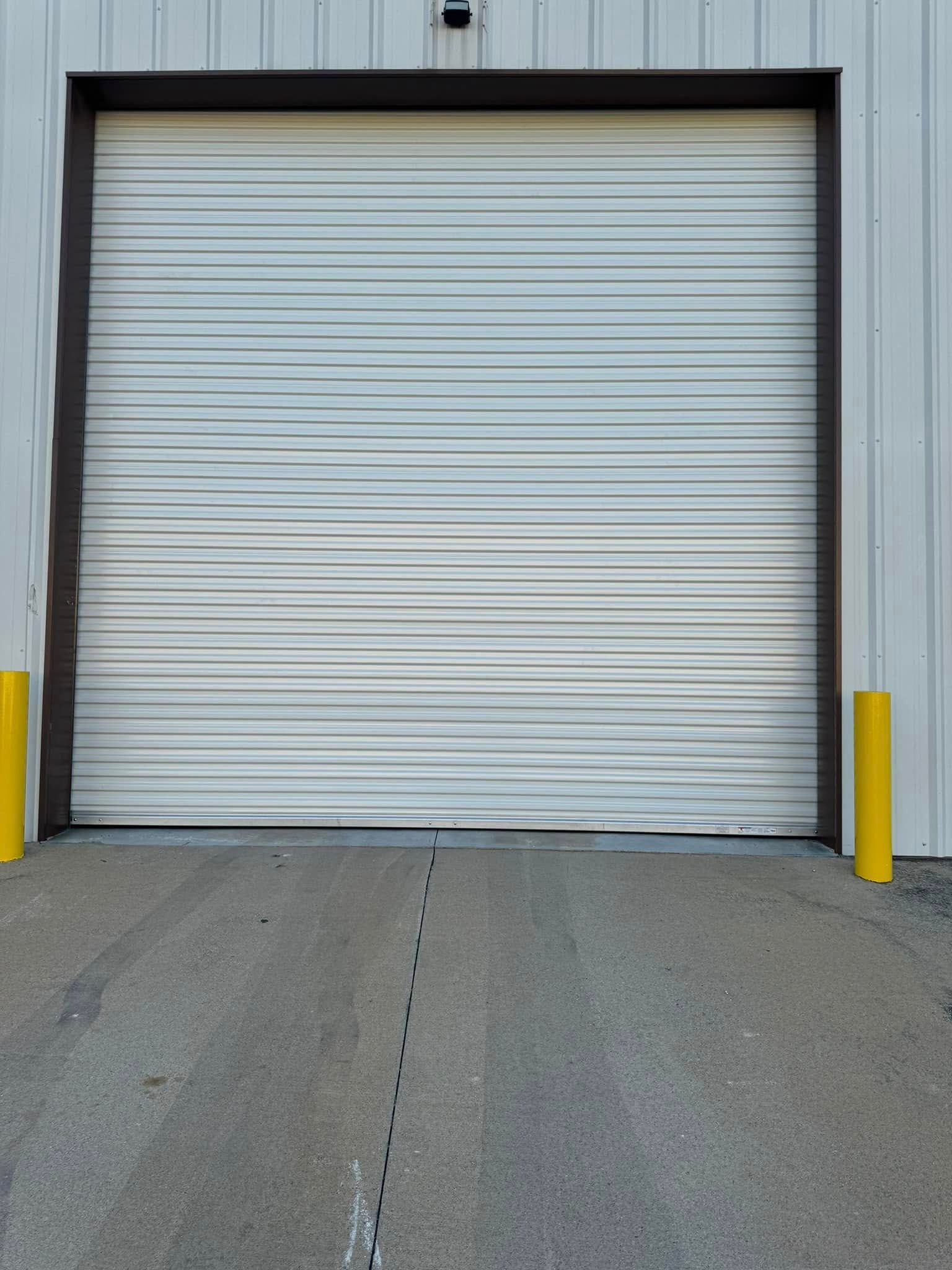Closed, light-colored roll-up garage door in a metal building, with yellow bollards on either side.