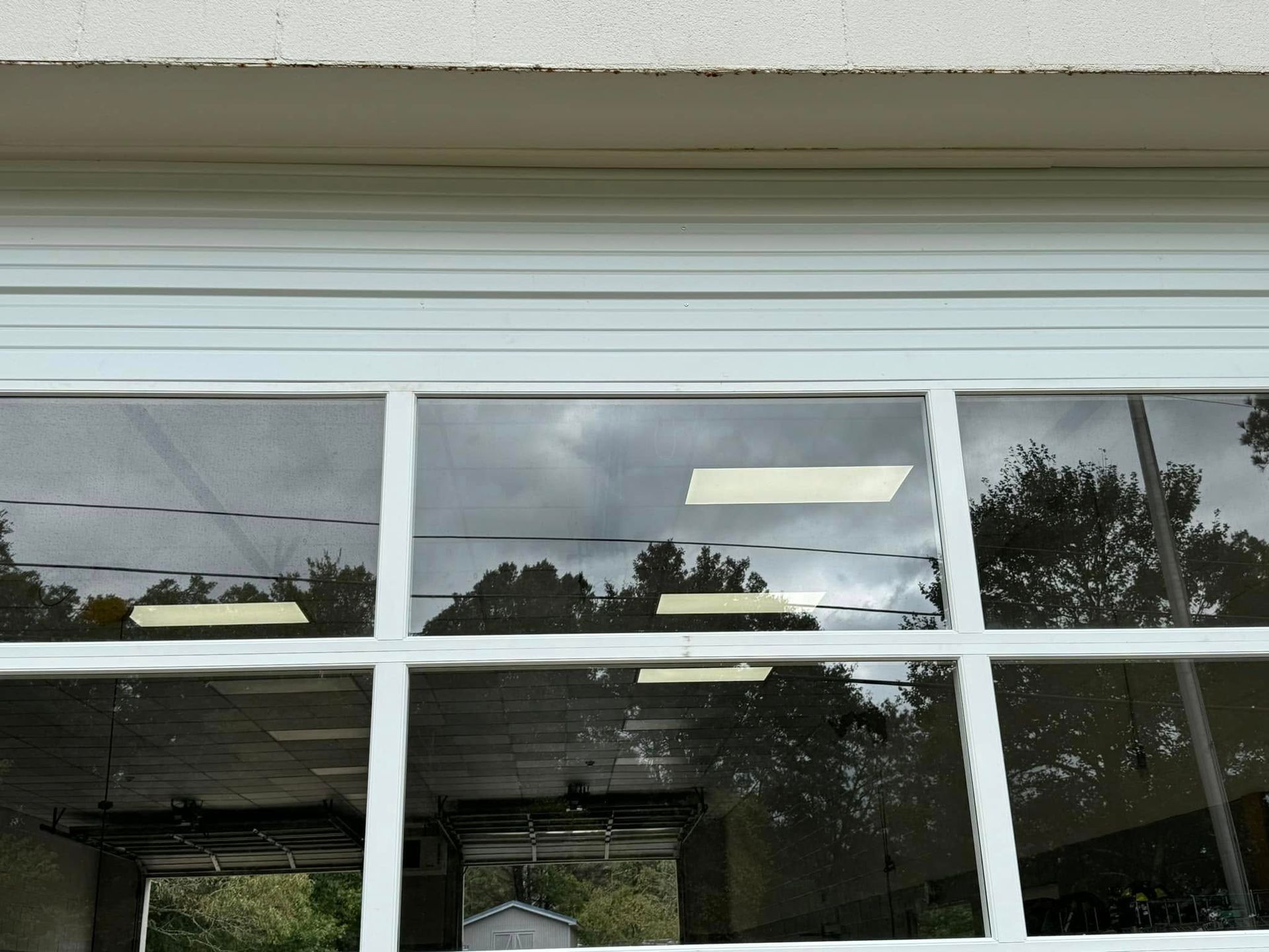 White building windows with a partially lowered shutter, reflecting trees and a cloudy sky.