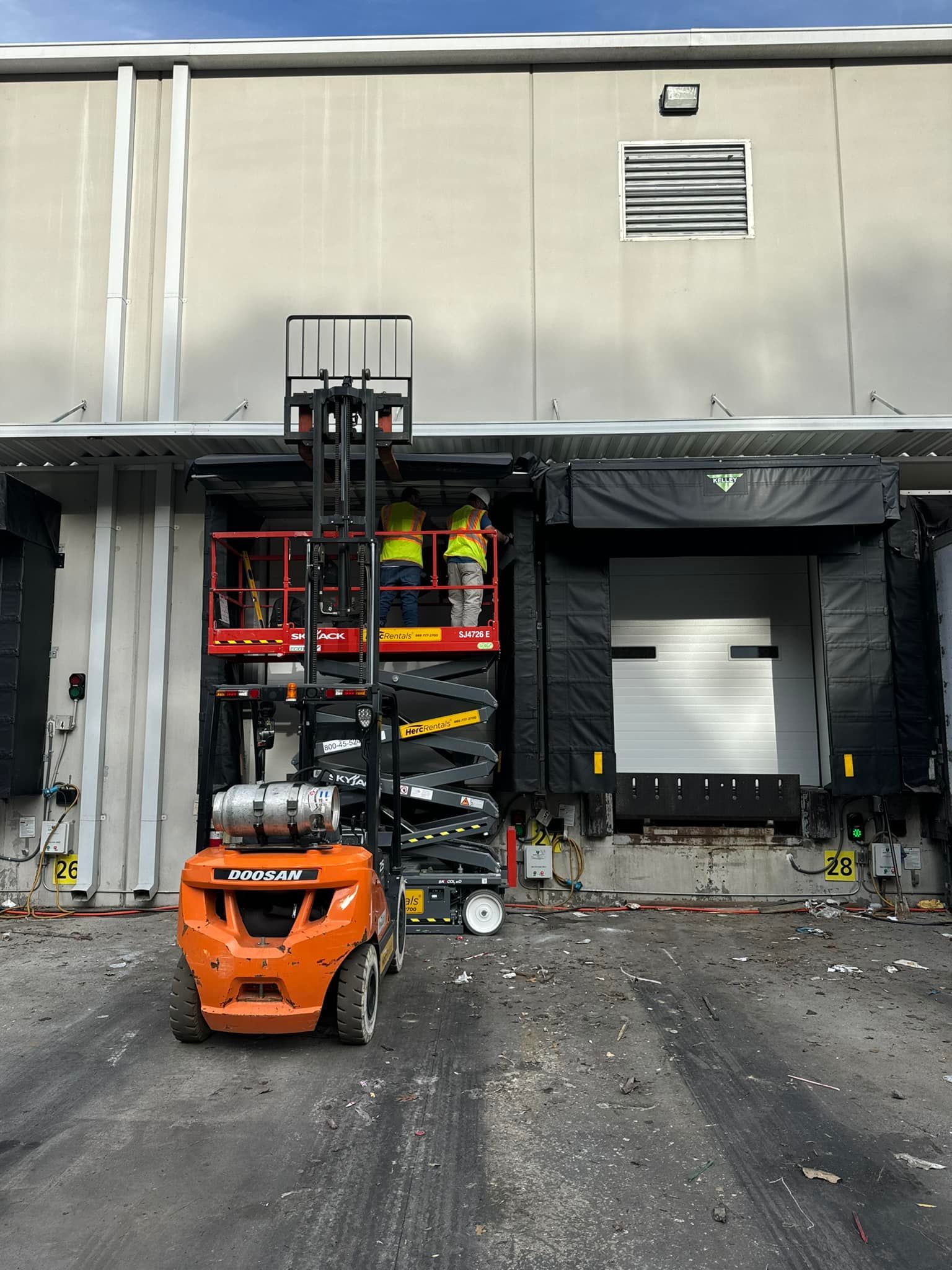Forklift with elevated platform at a loading dock; person in platform. Orange forklift, gray building, black dock seal.