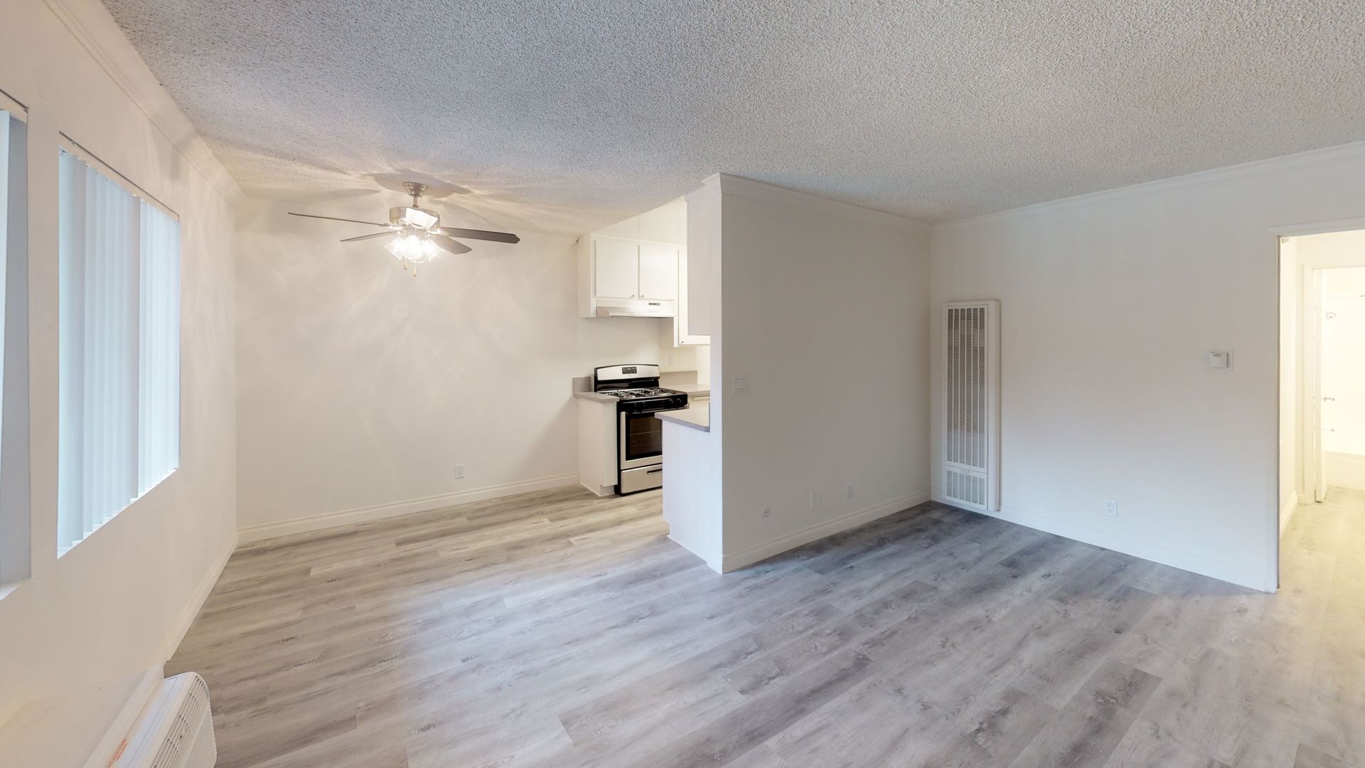 Interior of a small, bright apartment with white walls, gray wood-look flooring, and a small kitchen visible.