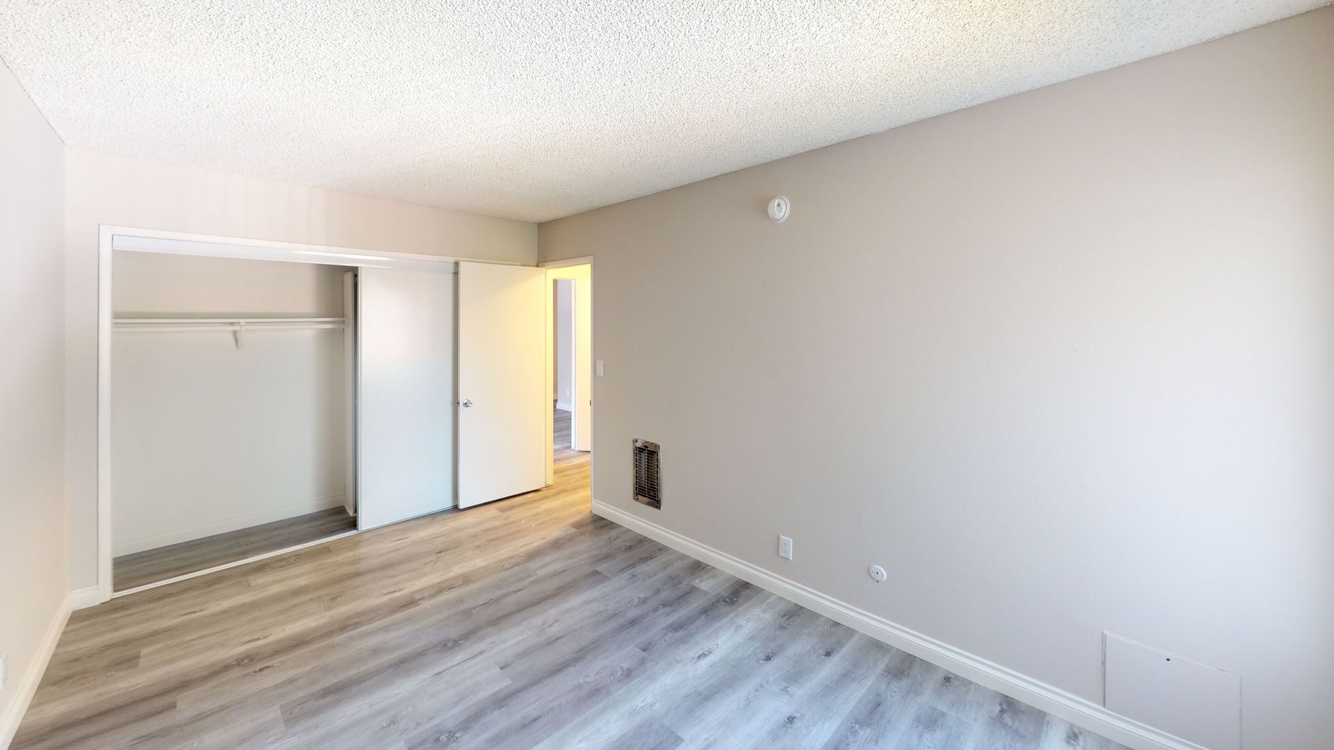 Empty bedroom with closet, light wood-look flooring, light grey walls, white doors, and a textured ceiling.