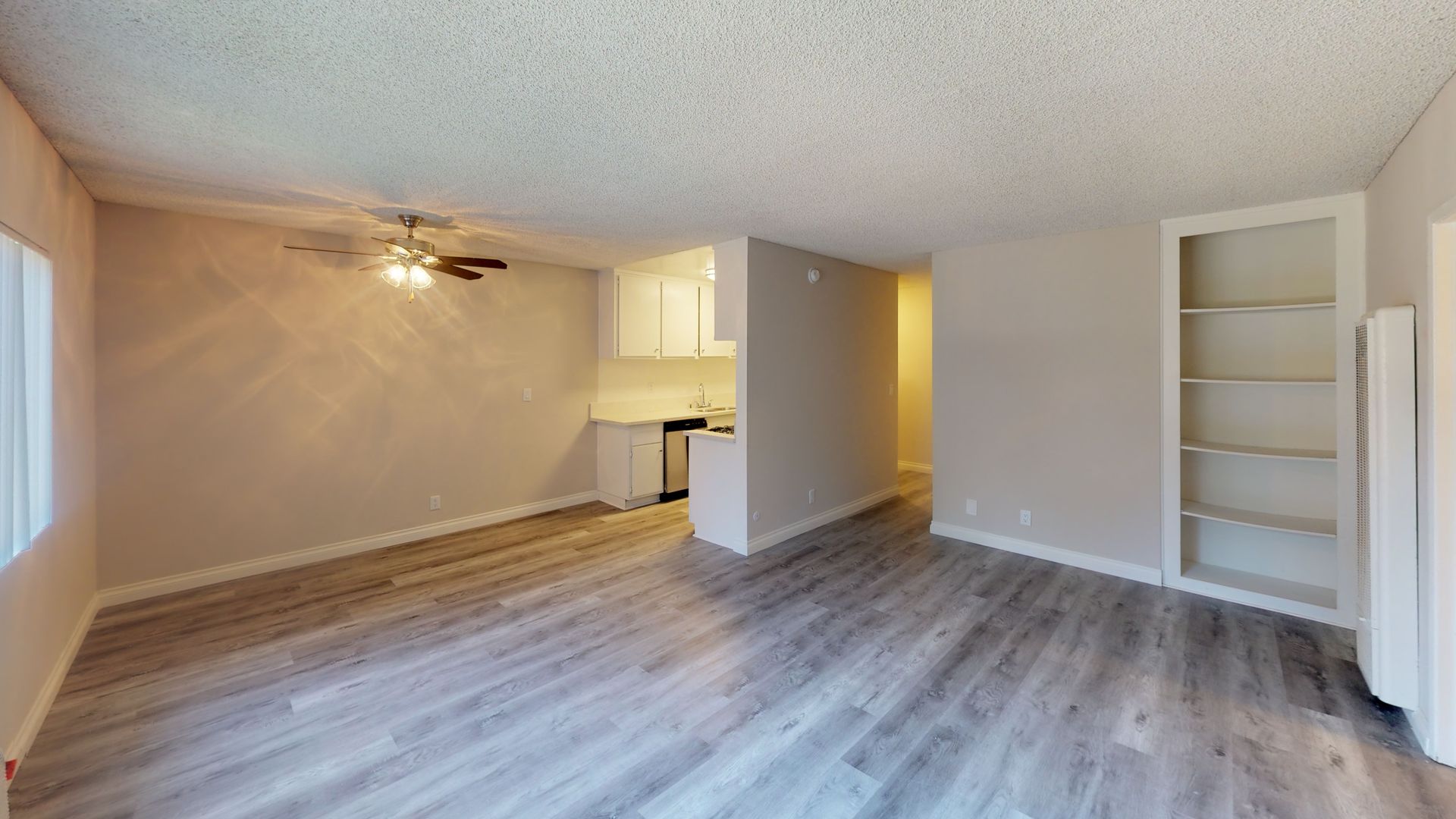 Empty living room with light grey wood-look floors, small kitchen, and built-in shelves.