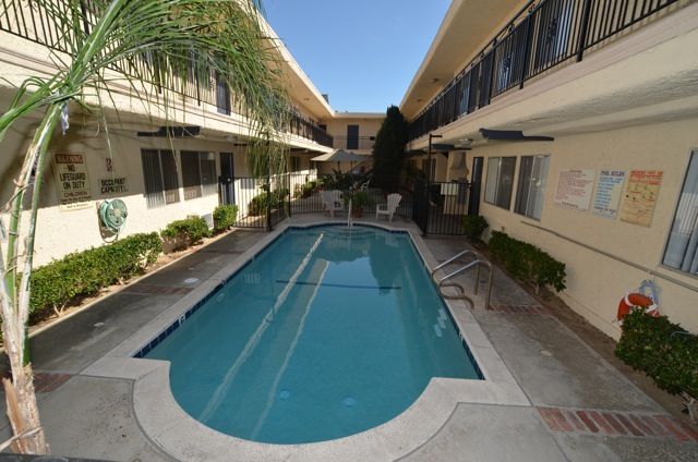Apartment complex courtyard with a rectangular swimming pool. Beige buildings, green bushes, and a blue sky.
