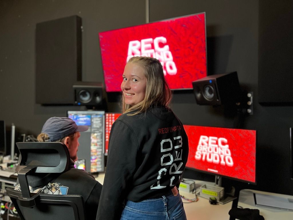 Eine Frau steht vor einem Mann, der in einem Aufnahmestudio an einem Schreibtisch sitzt.