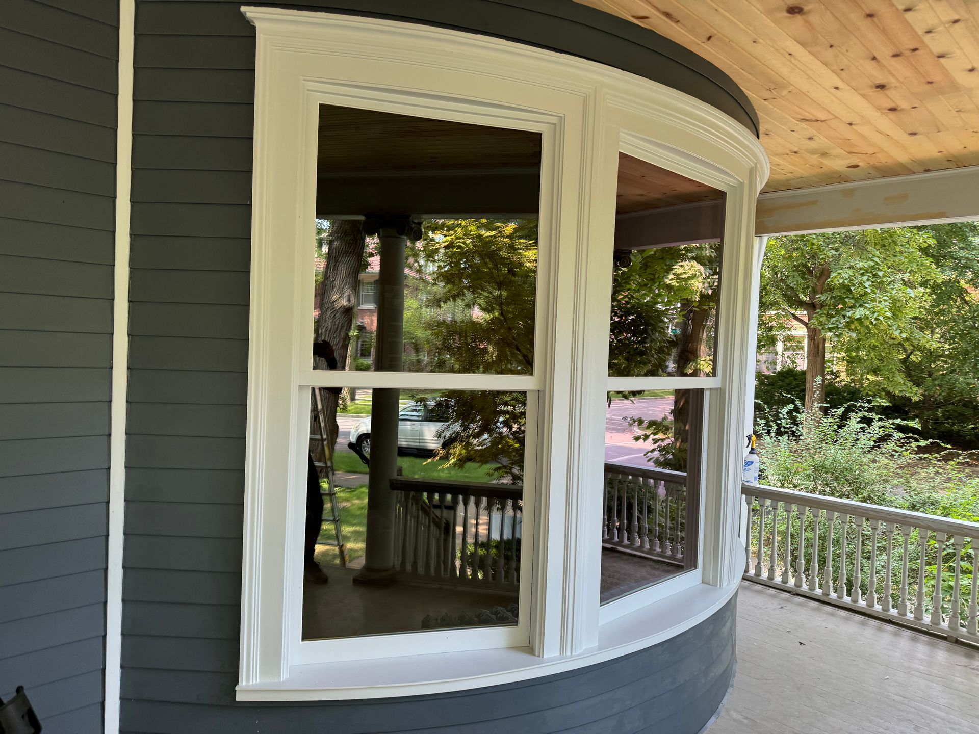 Curved window on gray house exterior, white trim, reflecting porch and trees.