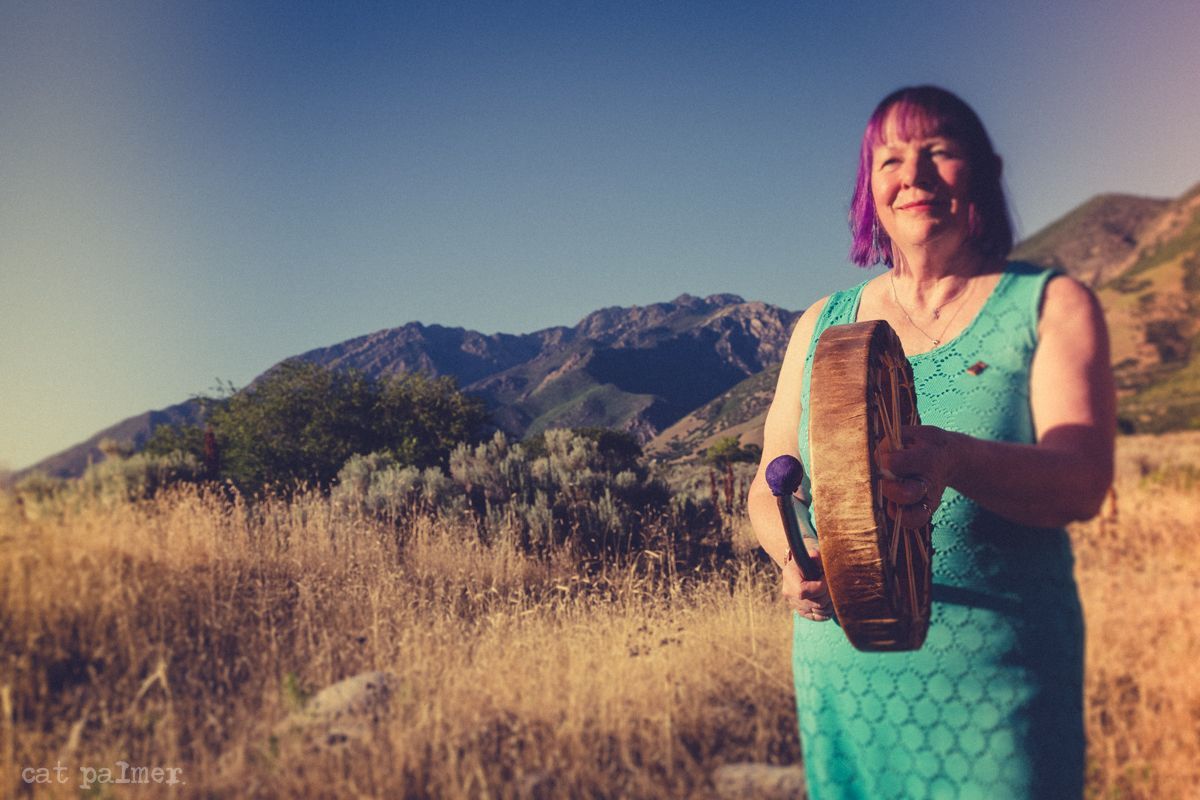 Woman with purple hair in a turquoise dress plays a drum in a field with mountains in the background.