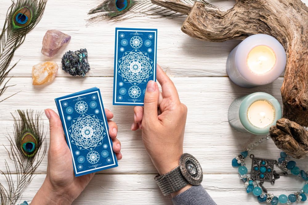Hands holding tarot cards with a blue decorative back, next to crystals, candles, and peacock feathers on wood.