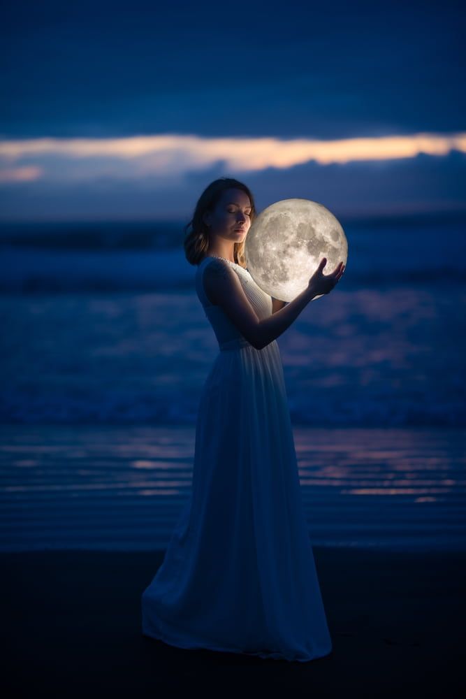 Woman holding illuminated moon sphere on a beach at dusk.