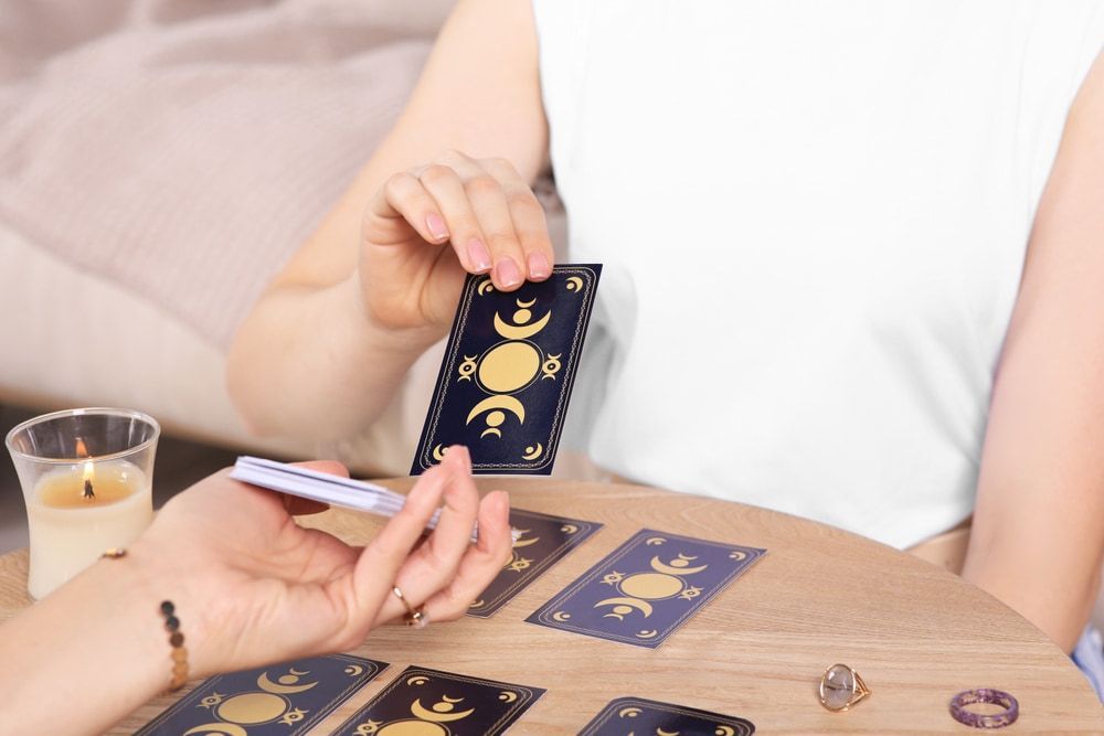 Woman draws a tarot card from a deck, laid on a table. Candle, other cards, and jewelry are also present.