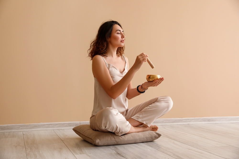 Woman in loungewear seated in meditation, striking a singing bowl.