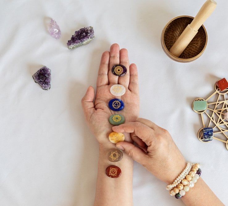 A hand with chakra stones, next to amethyst crystals, a singing bowl, and a geometric shape, on a white cloth.
