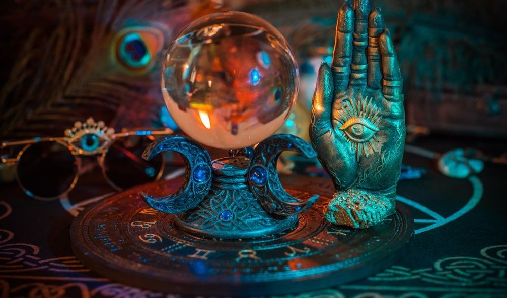 Crystal ball on crescent moon stand with open hand sculpture, peacock feather, and glasses.