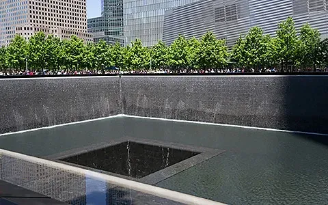 Two reflecting pools with cascading water, part of the 9/11 Memorial. Tall buildings and green trees are visible.