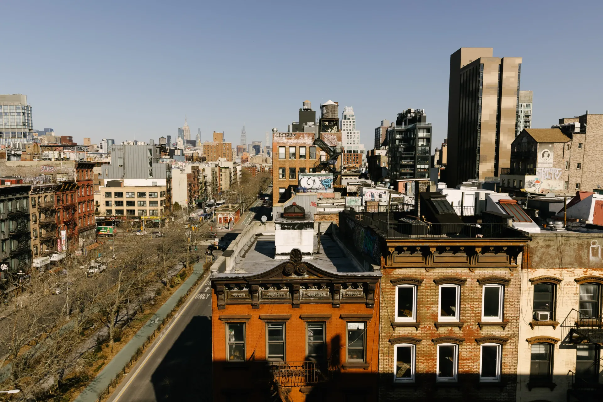 Cityscape with brownstone buildings, highway, and taller buildings in the distance, under a blue sky.