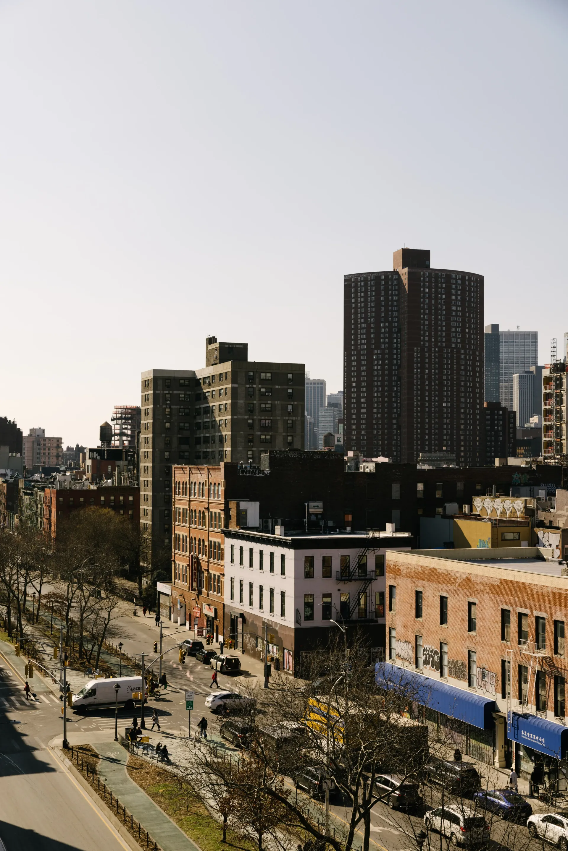 View of a city street lined with buildings, some tall. Cars and trees are along the street under a clear sky.